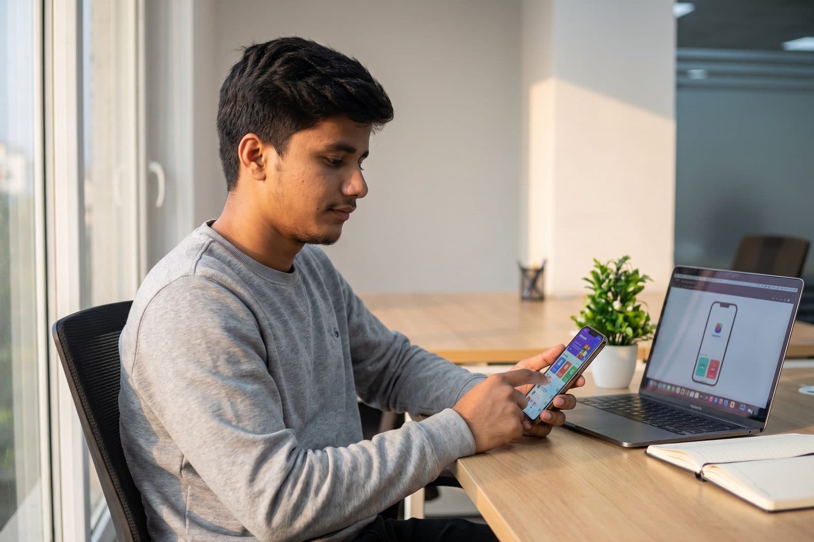 Manan Haider at a desk testing a mobile app on a phone, with the app preview open on a laptop beside him